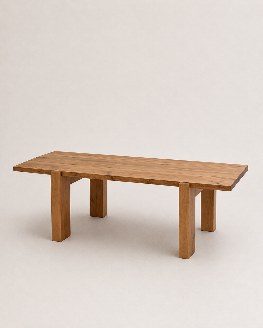 A minimalist rectangular wooden table with a natural oak finish, featuring thick block legs and a slightly tapered top, shown against a plain light gray studio background.