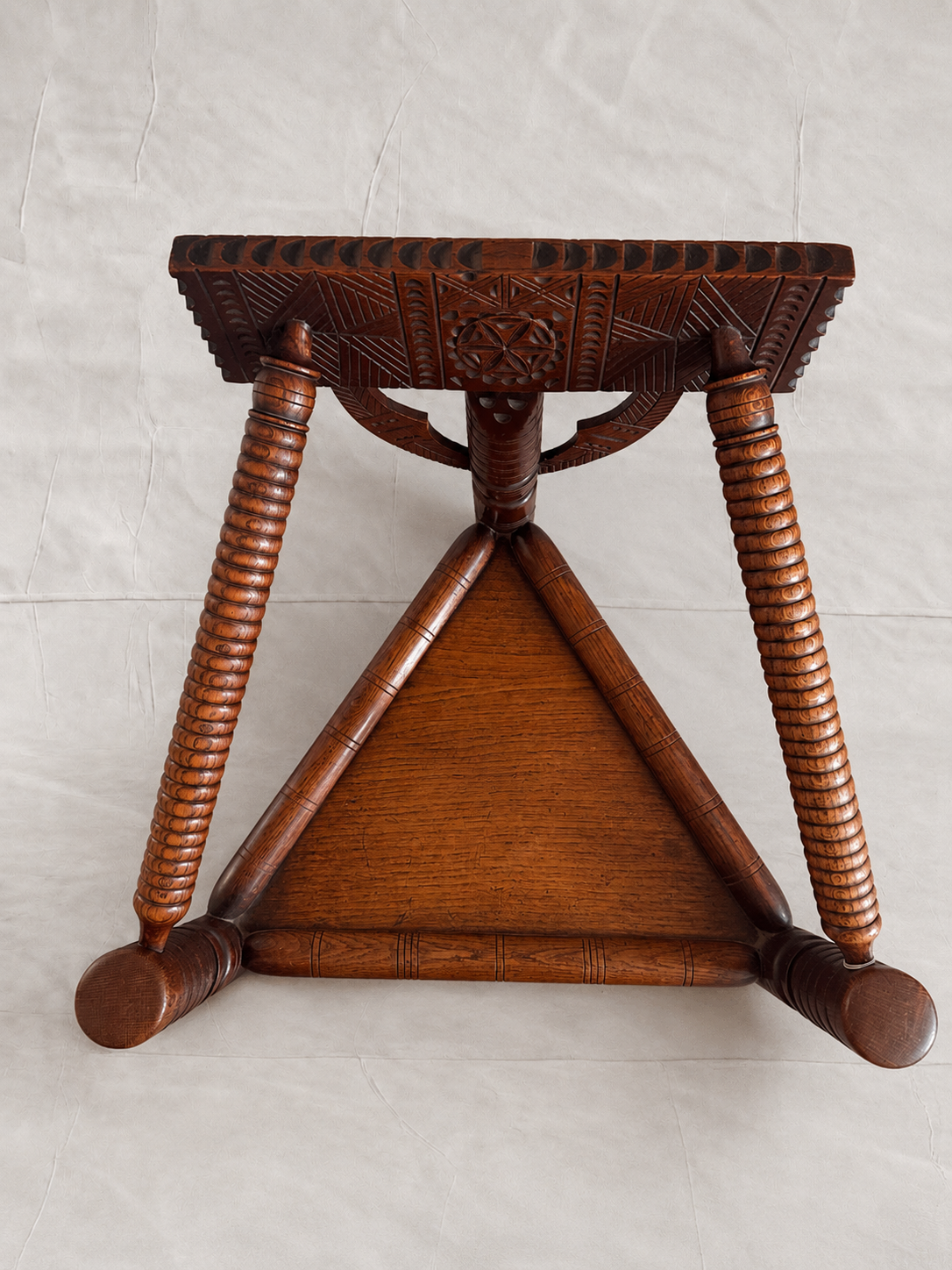 A dark brown carved wooden stool with a triangular base, twisted legs, and a patterned seat, photographed against a light gray fabric backdrop.