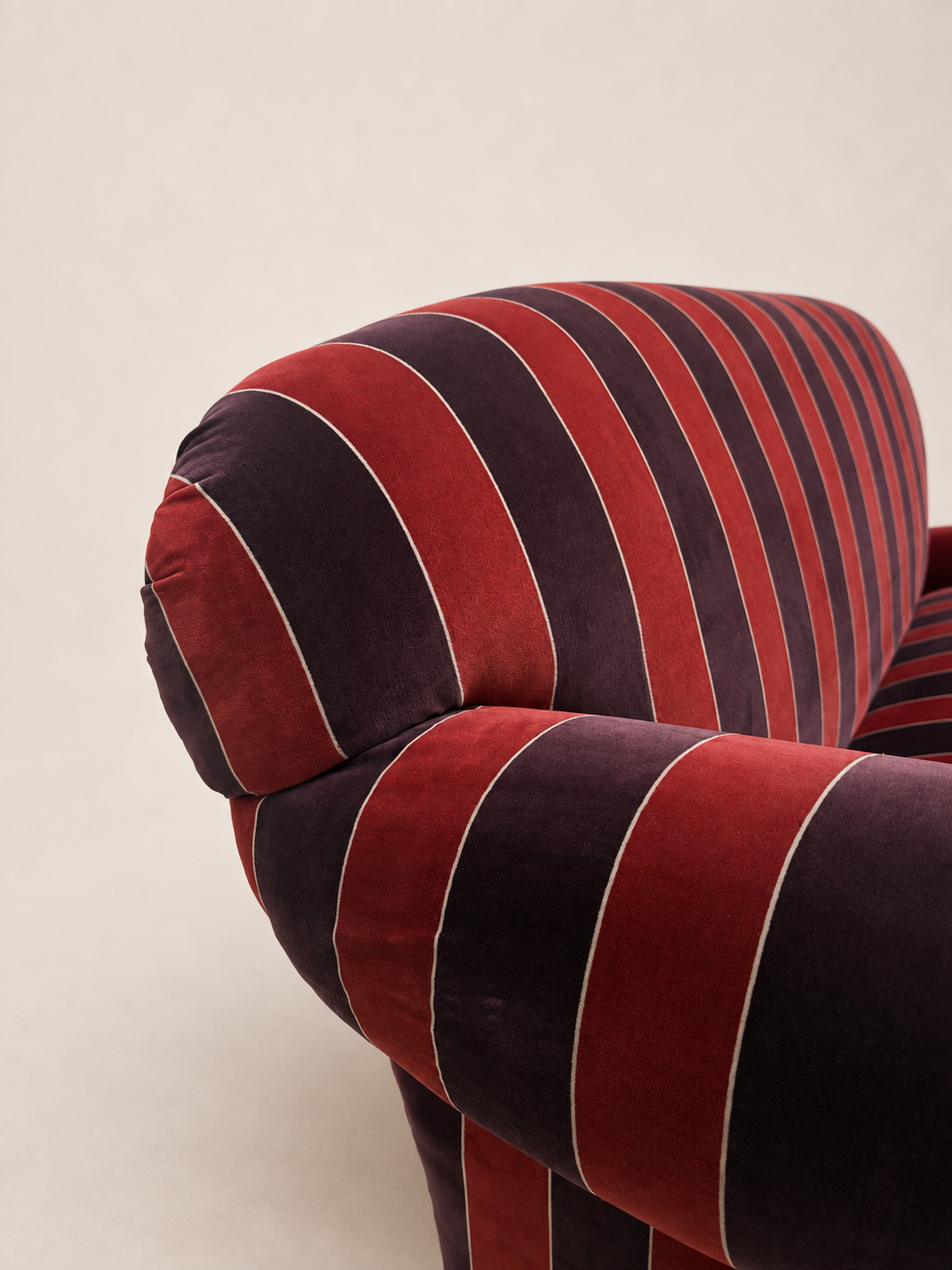 Close-up of a striped upholstered armchair in deep red and dark plum velvet with thin light piping, shown against a plain light beige background.