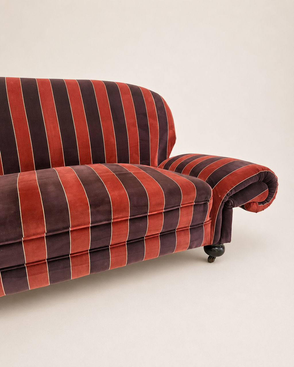 A close-up of a velvet sofa with vertical stripes in rust red, dark brown, and thin cream lines, shown against a plain off-white background.