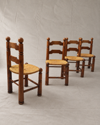 Four rustic Charles Dudouyt wooden dining chairs with woven straw seats, arranged on a light gray fabric backdrop.
