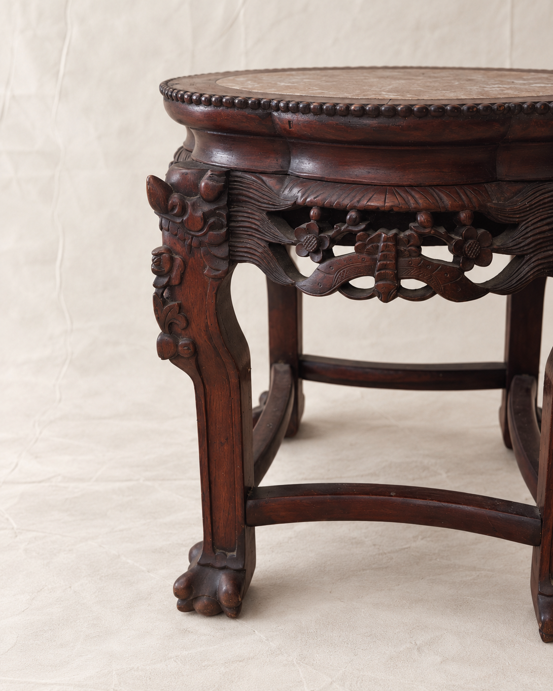 An antique dark brown carved wooden stool with ornate floral details on the legs and a circular seat, photographed against a neutral beige fabric backdrop.