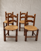 Four rustic Charles Dudouyt wooden children's chairs with woven straw seats, arranged in a group against a neutral gray fabric backdrop.