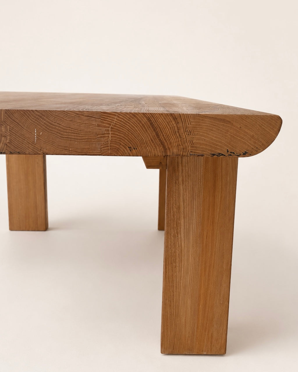 Close-up of a rustic wooden table leg and tabletop corner, showing natural oak wood grain and a curved apron detail against a plain off-white background.