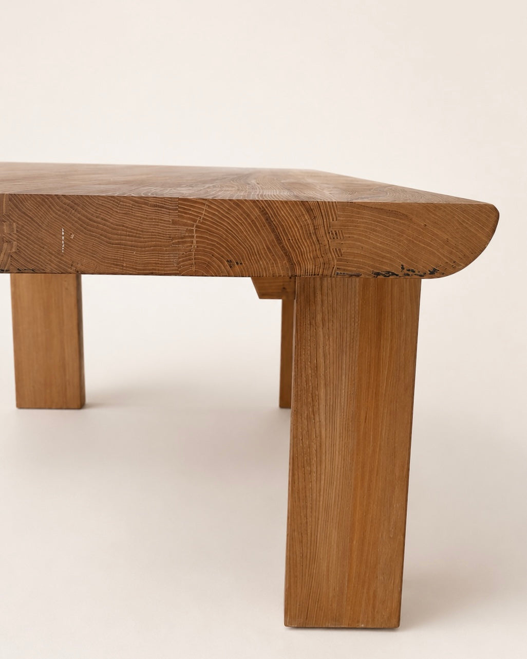 Close-up of a rustic wooden table leg and tabletop corner, showing natural oak wood grain and a curved apron detail against a plain off-white background.