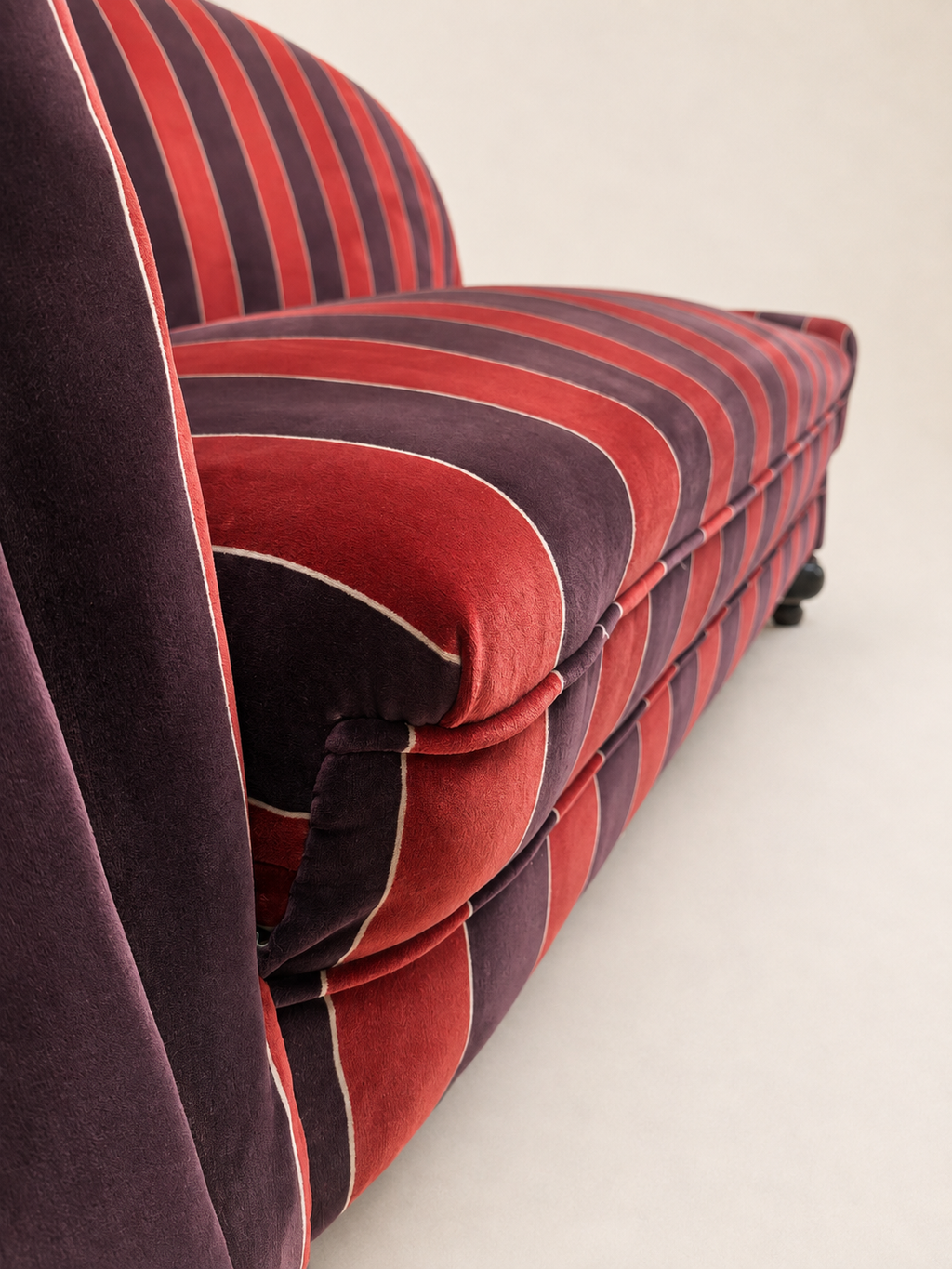 A close-up of an upholstered armchair or sofa corner with bold vertical stripes in red, dark plum velvet, showing the seat cushion, backrest, and side against a plain light background.