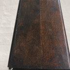 A close-up of a dark brown wooden surface, likely a table top or cabinet panel, showing visible grain and wear, photographed on a light beige tiled floor.