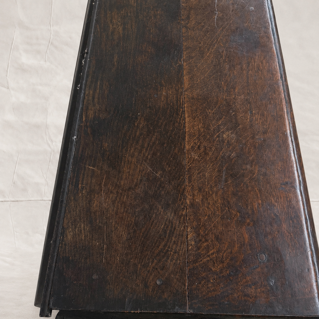 A close-up of a dark brown wooden surface, likely a table top or cabinet panel, showing visible grain and wear, photographed on a light beige tiled floor.