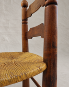 A close-up of a rustic Charles Dudouyt wooden chair with a woven straw seat, showing the dark brown wood frame and the natural fiber texture against a neutral light gray backdrop.