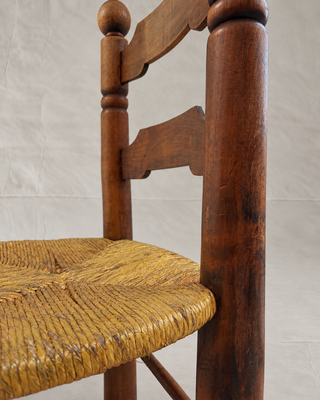 A close-up of a rustic Charles Dudouyt wooden chair with a woven straw seat, showing the dark brown wood frame and the natural fiber texture against a neutral light gray backdrop.