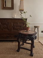 A dark carved wooden side table with a round stone-top surface stands in a styled room setting next to a large wooden cabinet, with a small potted plant on top.
