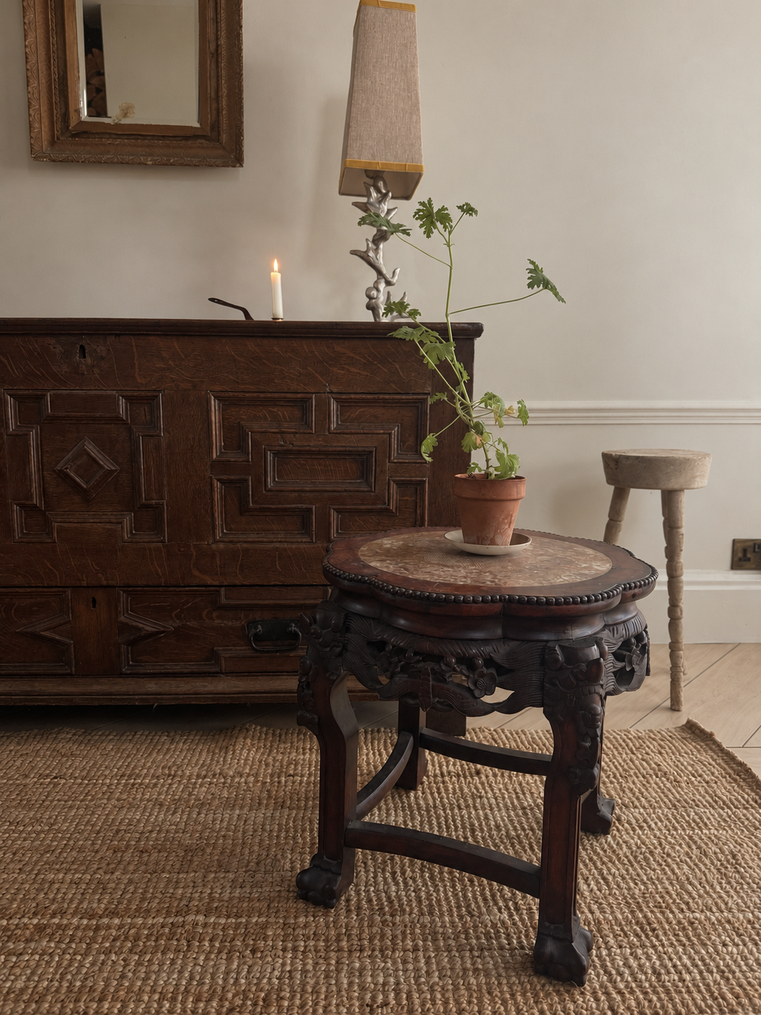 A dark carved wooden side table with a round stone-top surface stands in a styled room setting next to a large wooden cabinet, with a small potted plant on top.