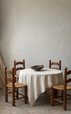 A round dining table covered with a white linen tablecloth, surrounded by four wooden chairs with woven seats, set against a textured plaster wall.
