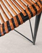 Close-up detail of a chair seat with curved brown leather straps wrapped around a black metal frame, set against a light beige textured floor.