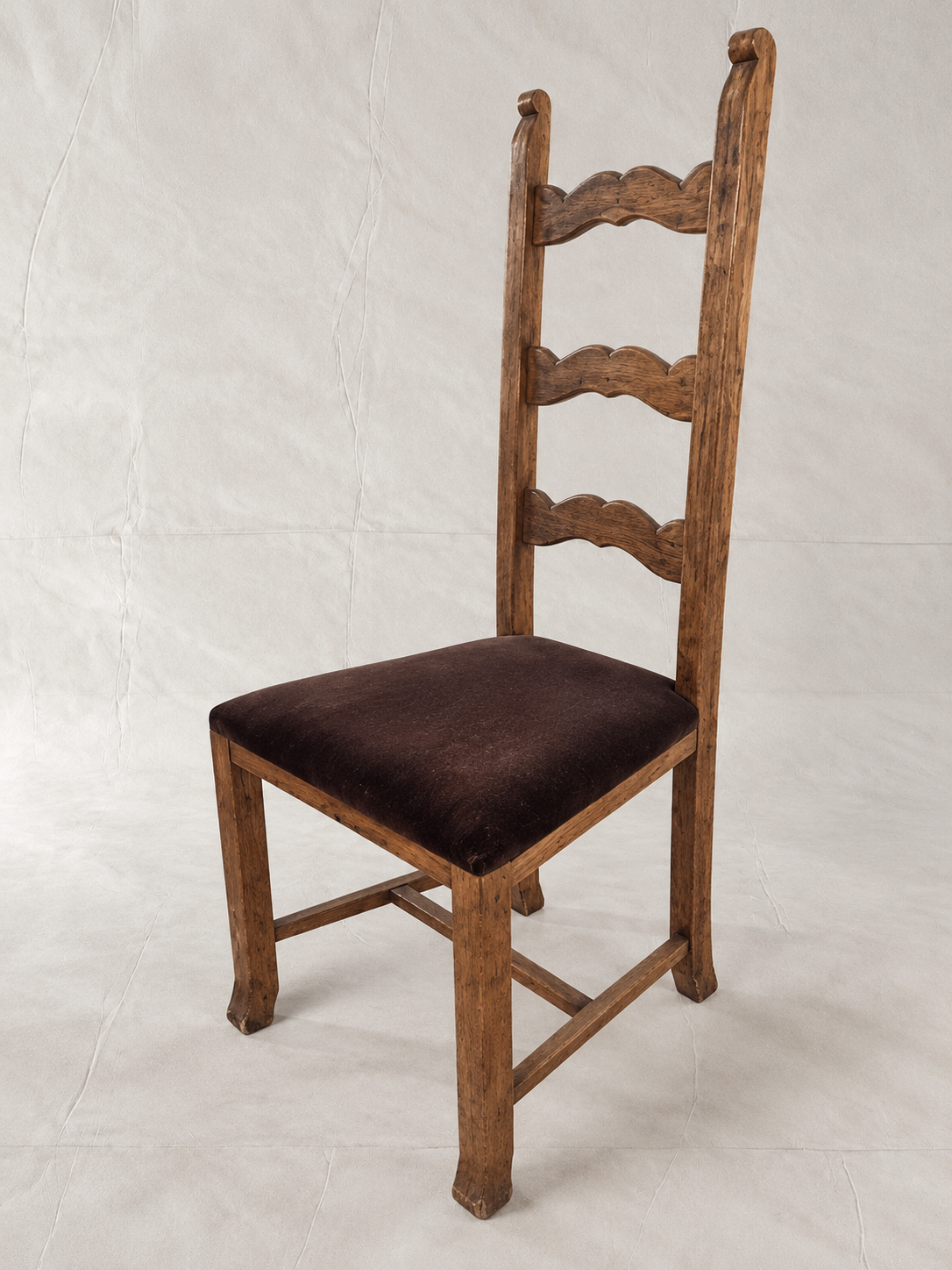 A wooden ladder-back dining chair with a dark brown upholstered seat, photographed against a wrinkled off-white studio backdrop.
