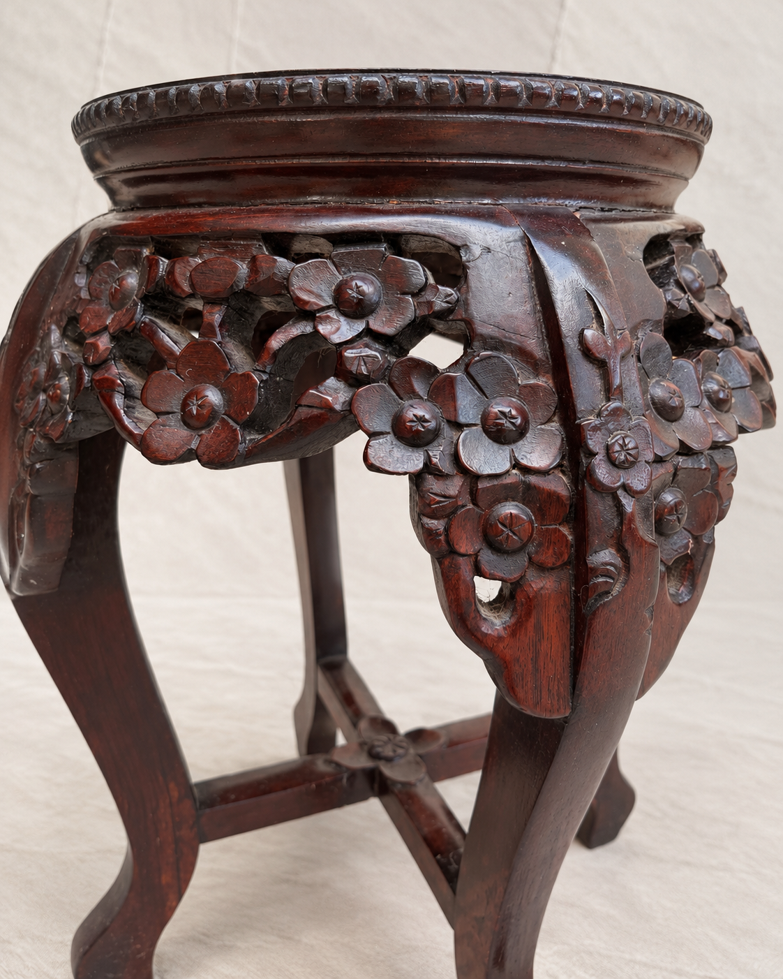 An ornate dark reddish-brown wooden stand or stool with carved floral details on the sides, photographed against a light neutral fabric backdrop.