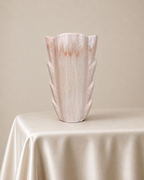 A sculptural ceramic vase with a flared, layered leaf-like silhouette, finished in a distressed light pink and cream glaze, displayed on a draped cream satin tablecloth against a solid beige background.