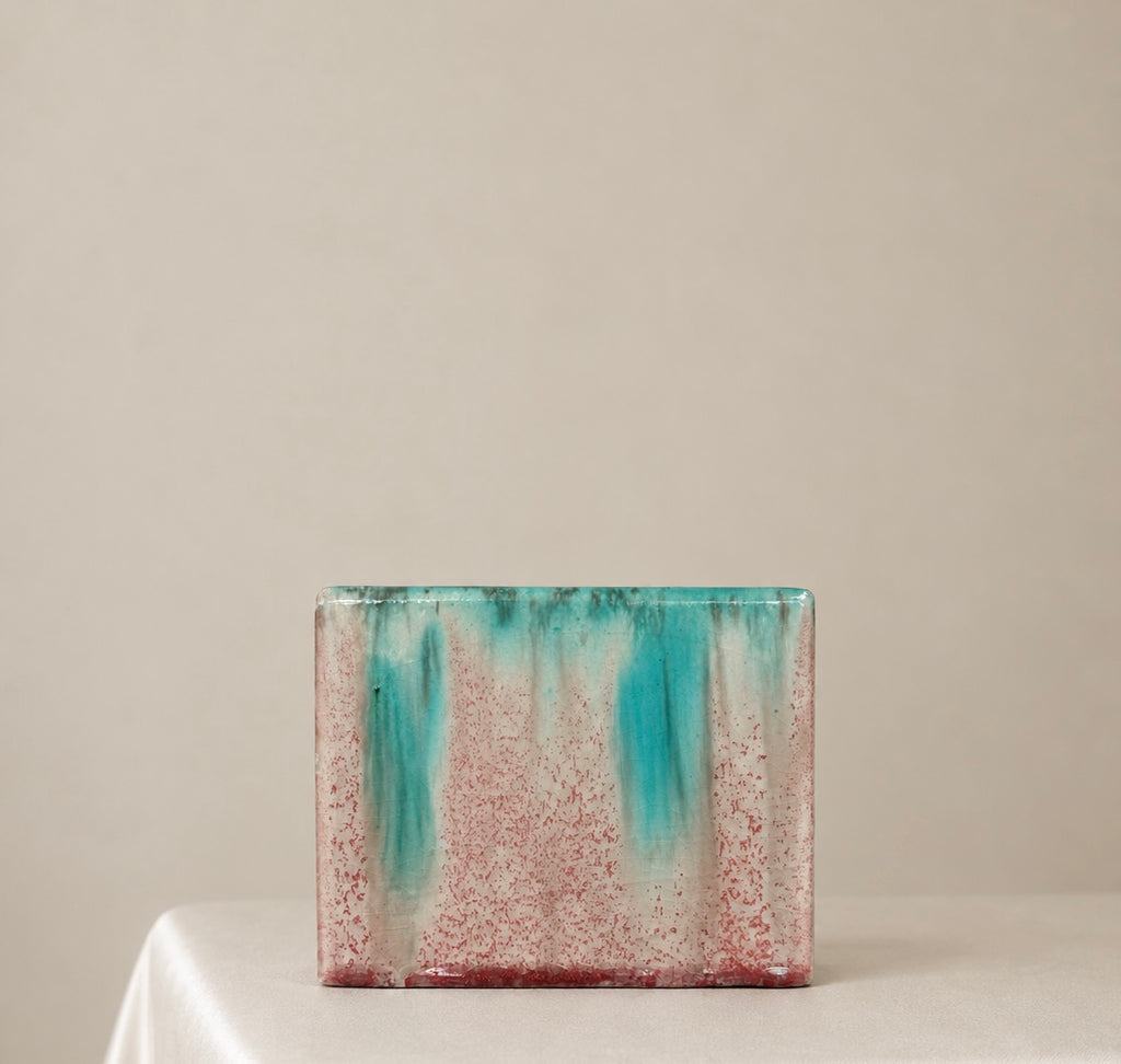 A rectangular ceramic vase with a textured, speckled glaze in pink and turquoise, sitting on a white draped table against a neutral beige background.