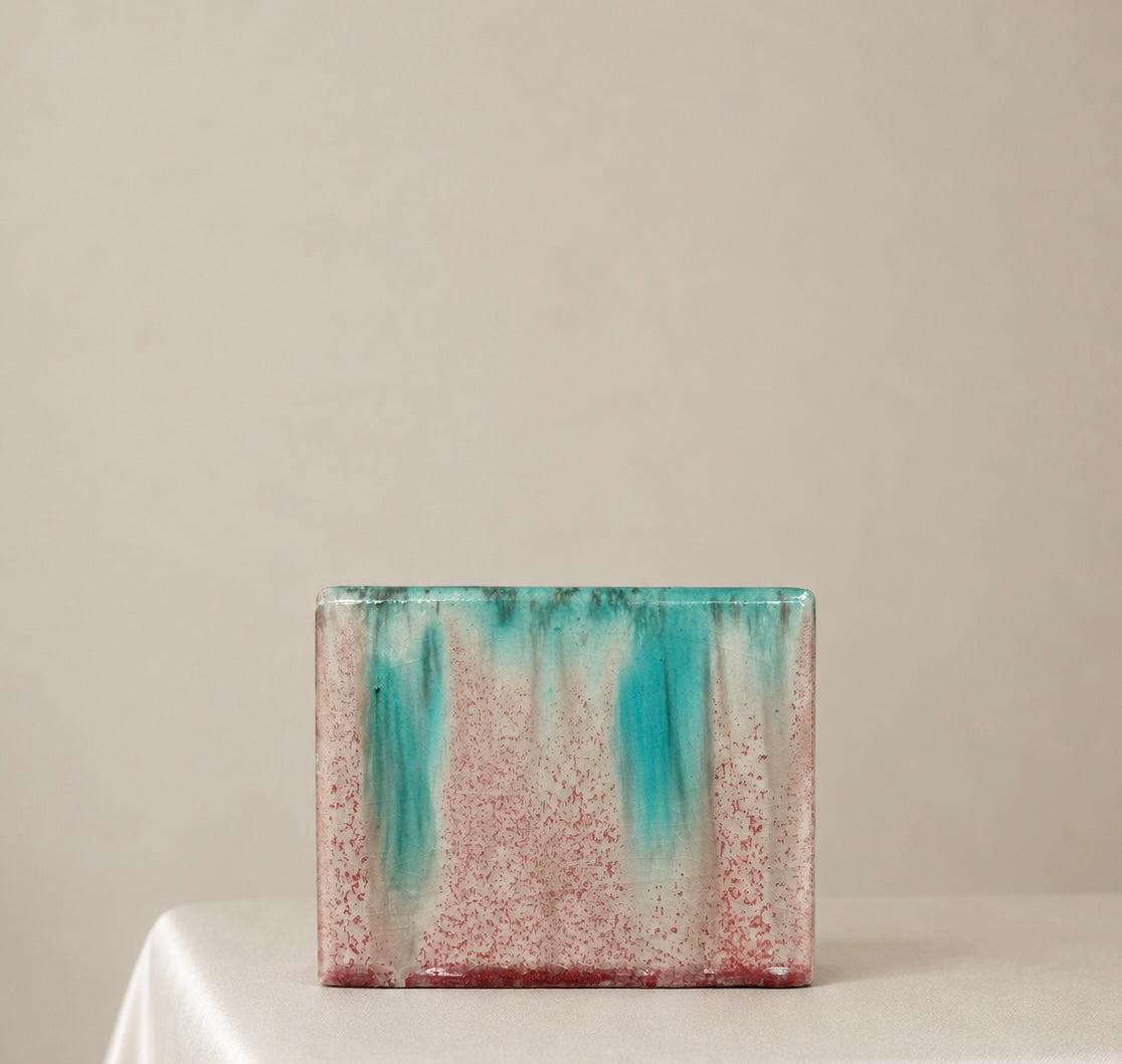 A rectangular ceramic vase with a textured, speckled glaze in pink and turquoise, sitting on a white draped table against a neutral beige background.