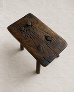 A rustic dark brown wooden stool with a thick rectangular seat, four legs, and four round metal pegs on top, photographed from an elevated angle on a light beige tiled floor.
