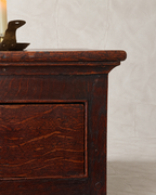 Close-up of a dark reddish-brown wooden chest or cabinet with a visible drawer front and natural wood grain, photographed against a light neutral wall.