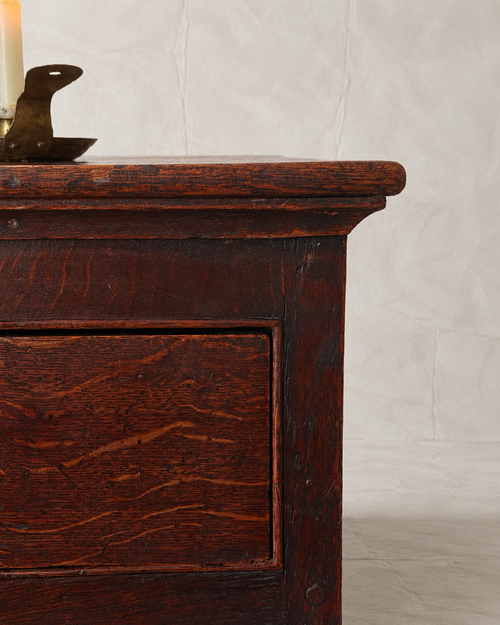Close-up of a dark reddish-brown wooden chest or cabinet with a visible drawer front and natural wood grain, photographed against a light neutral wall.