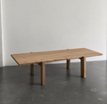 A rectangular wooden dining table with thick block legs, shown in a minimalist studio setting with a gray concrete floor and a light gray wall.