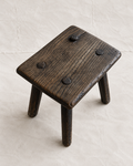 A rustic dark brown wooden stool with a square seat and three legs, featuring four dark metal rivets on the top surface, set against a light beige textured background.