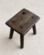 A rustic dark brown wooden stool with a square seat and three legs, featuring four dark metal rivets on the top surface, set against a light beige textured background.