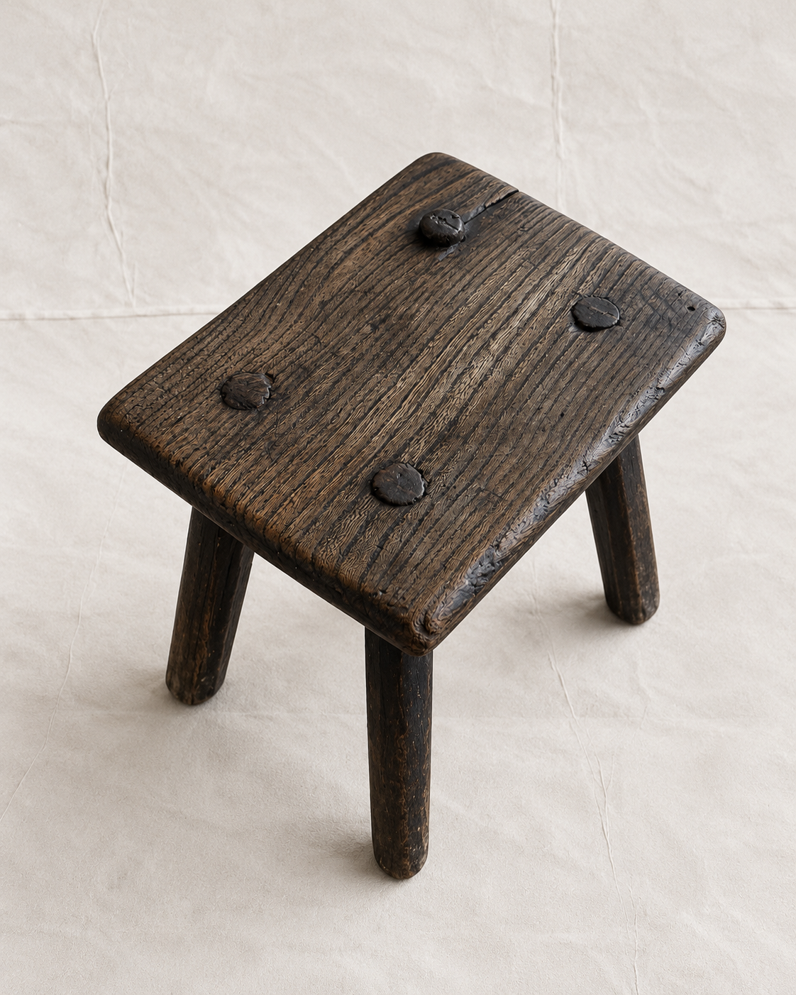 A rustic dark brown wooden stool with a square seat and three legs, featuring four dark metal rivets on the top surface, set against a light beige textured background.