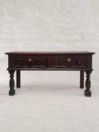 A dark brown wooden console table with two drawers, turned legs, and brass hardware, photographed against a wrinkled off-white backdrop.