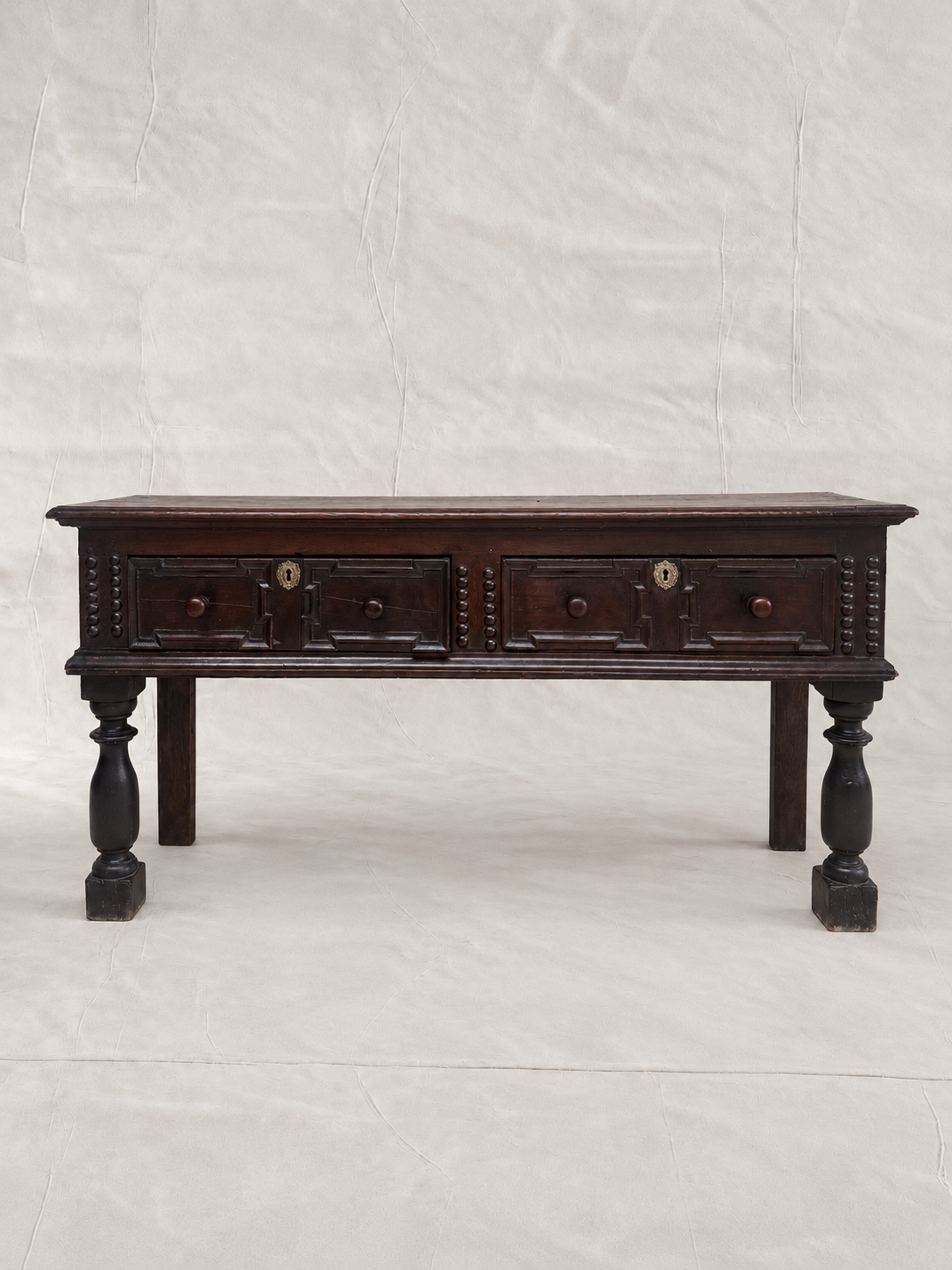 A dark brown wooden console table with two drawers, turned legs, and brass hardware, photographed against a wrinkled off-white backdrop.