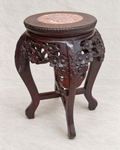 A dark reddish-brown carved wooden stool with floral details on the sides and a round pink stone top, photographed against a light fabric backdrop.