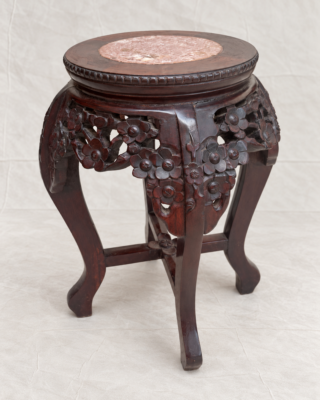 A dark reddish-brown carved wooden stool with floral details on the sides and a round pink stone top, photographed against a light fabric backdrop.