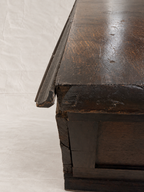Close-up of a dark stained wooden table corner showing the thick apron, mitered edge, and worn wood grain against a white studio backdrop.