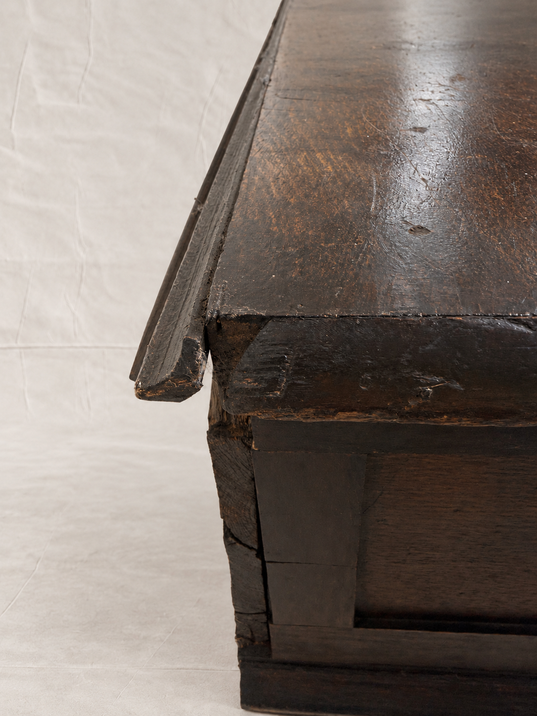 Close-up of a dark stained wooden table corner showing the thick apron, mitered edge, and worn wood grain against a white studio backdrop.
