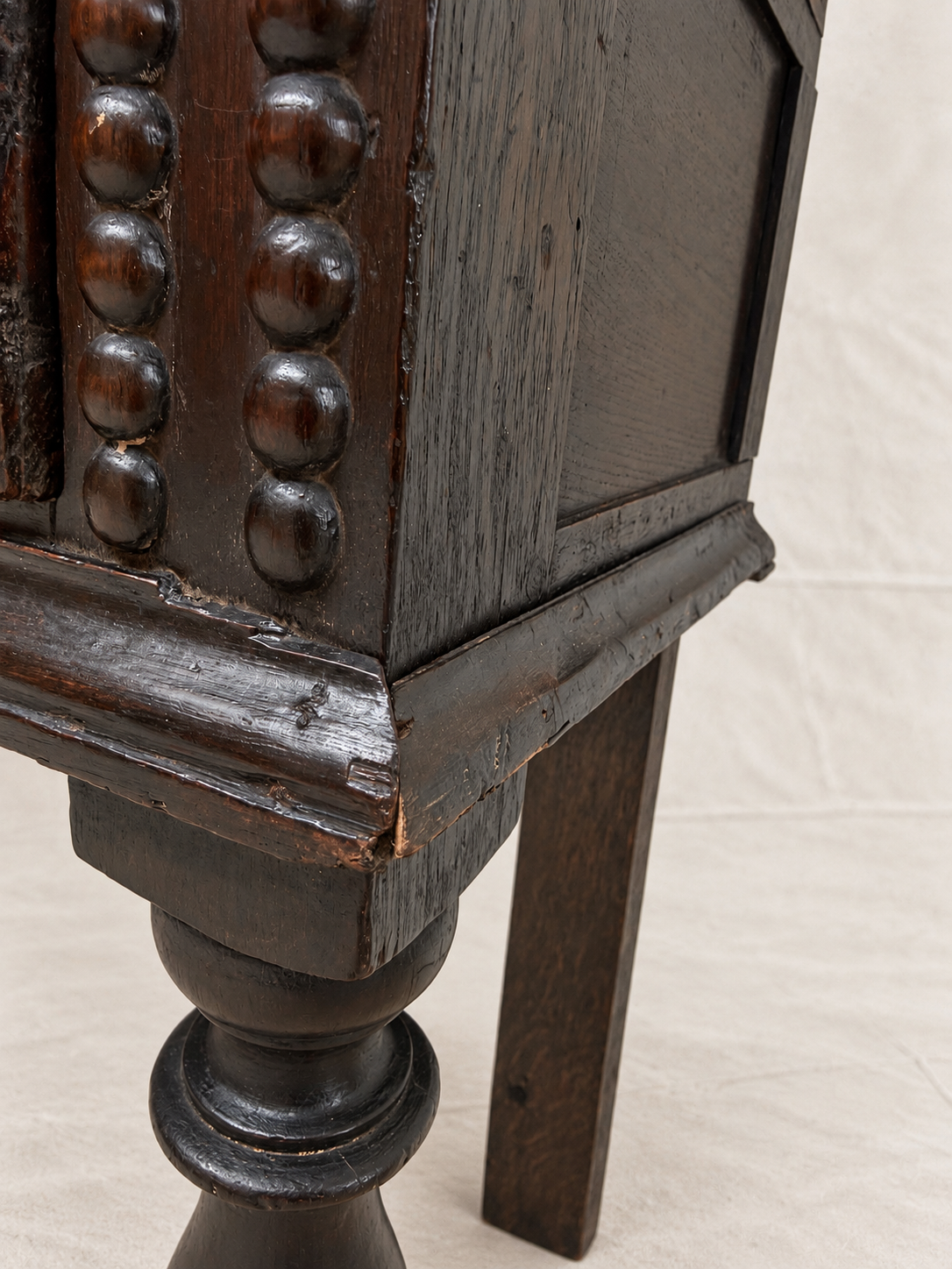 Close-up detail of a dark brown wooden cabinet or side table, showing carved beaded vertical panels, a turned leg, and a light off-white fabric background.