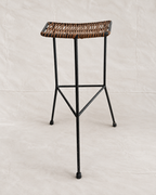 A modern bar stool with a black metal frame and a woven brown wicker seat, photographed against a neutral light beige fabric backdrop.