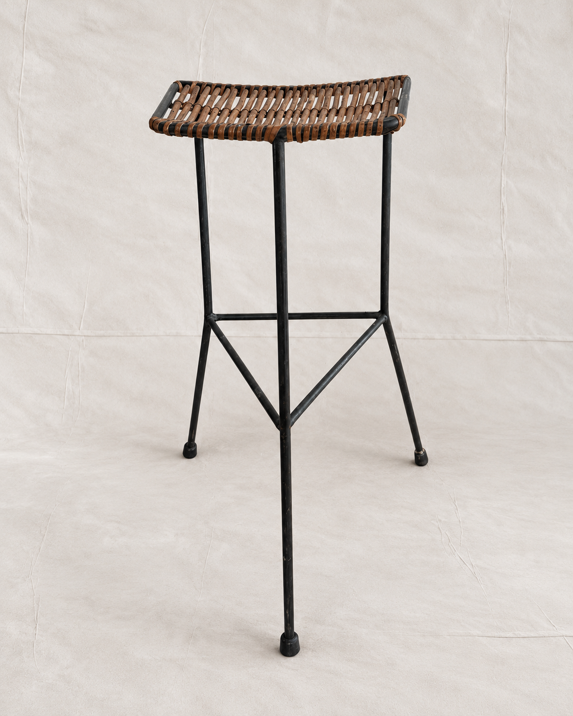 A modern bar stool with a black metal frame and a woven brown wicker seat, photographed against a neutral light beige fabric backdrop.