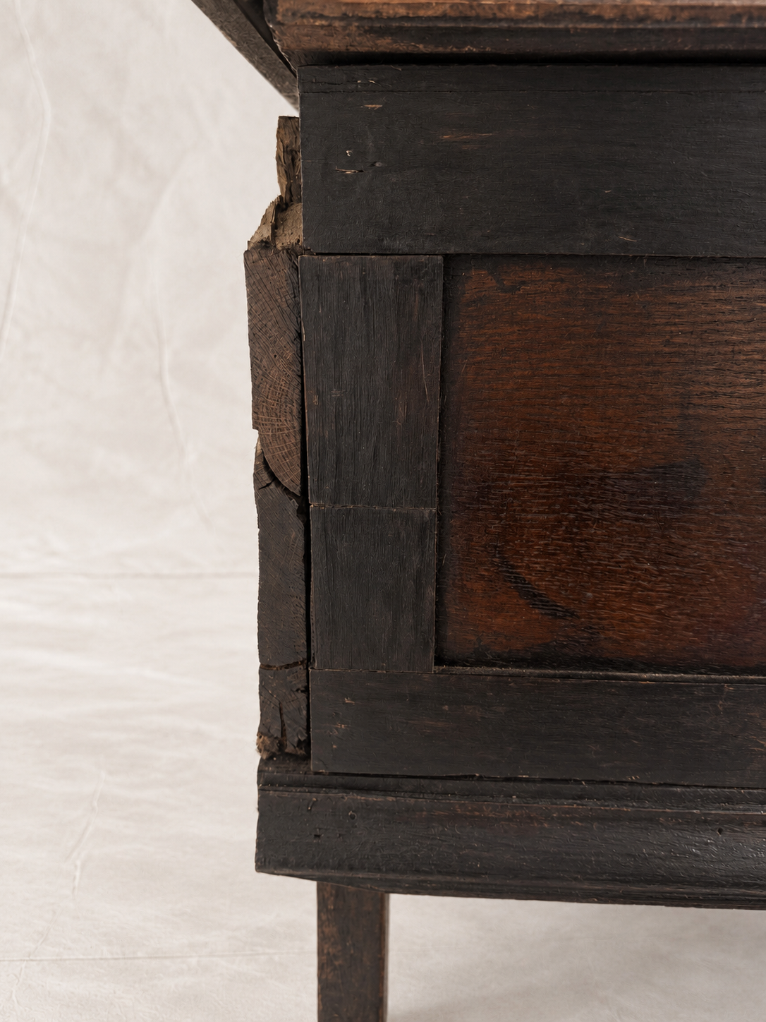 Close-up of a dark stained wooden furniture piece showing the side panel, front edge, and a visible crack in the wood, photographed against a white fabric backdrop.
