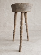 A rustic wooden stool with a round seat and three turned spindle legs, shown against a neutral textured beige backdrop.
