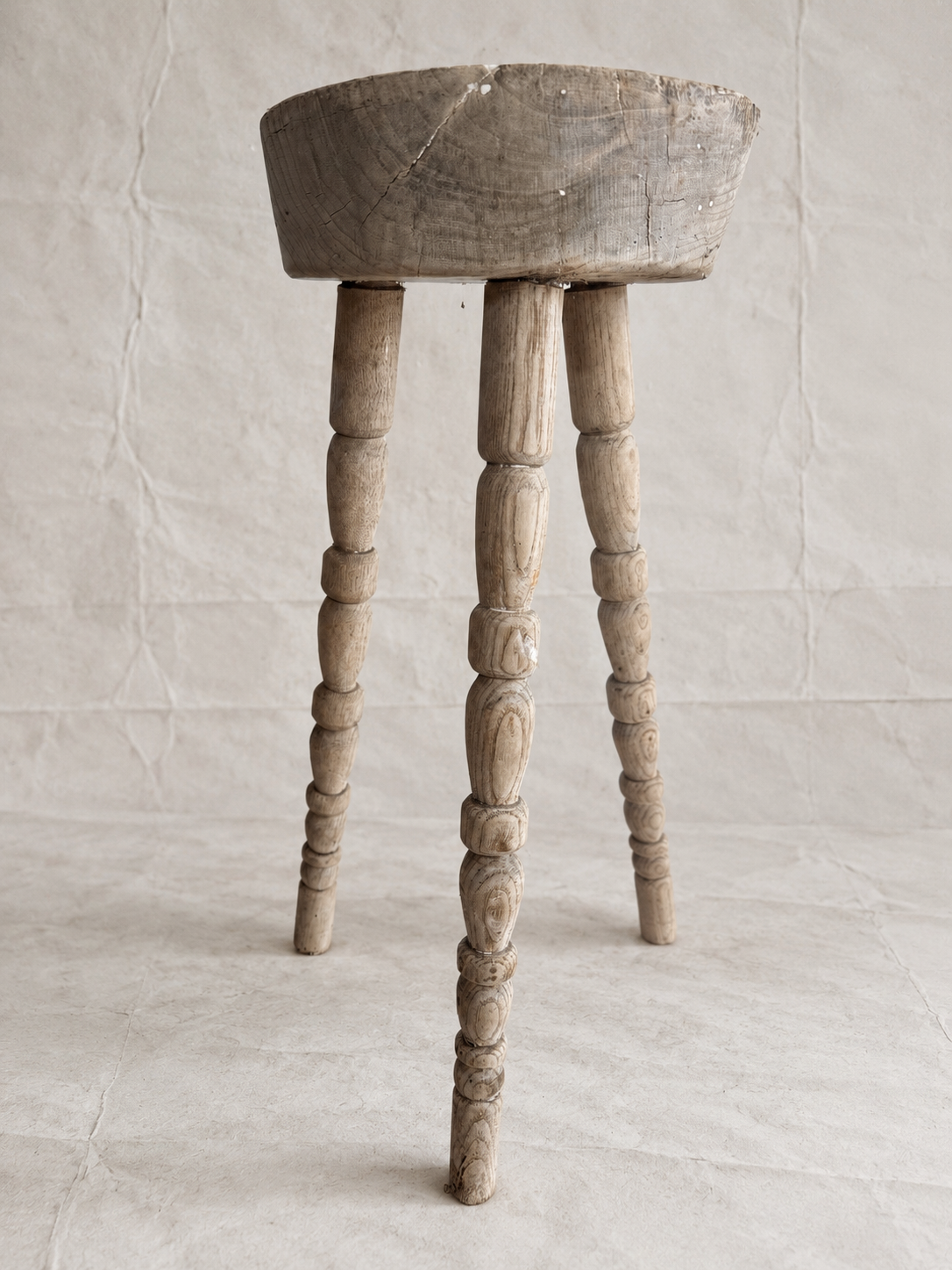 A rustic wooden stool with a round seat and three turned spindle legs, shown against a neutral textured beige backdrop.