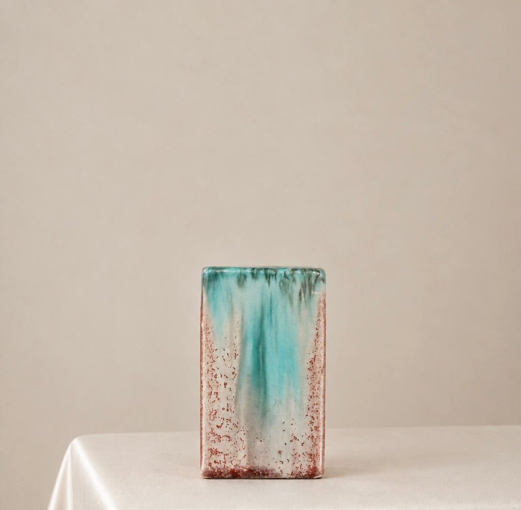 A rectangular ceramic vase with a distressed glazed finish, featuring a vertical gradient of turquoise, teal, and rust-brown on a white base, displayed on a draped white tablecloth against a neutral beige background.