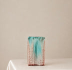 A rectangular ceramic vase with a distressed glazed finish, featuring a vertical gradient of turquoise, teal, and rust-brown on a white base, displayed on a draped white tablecloth against a neutral beige background.