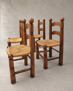 Four rustic Charles Dudouyt wooden dining chairs with woven straw seats and round wooden backrests, arranged on a light gray fabric backdrop.