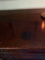 A close-up of a dark brown wooden dresser or chest with visible wear, scratches, and a dark stain on the top surface.