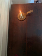 A lit white candle in a small round wooden holder with a handle, mounted on a dark wooden door or panel in an indoor setting.