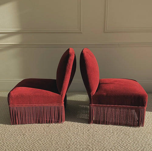 Two red velvet chairs with fringe on a beige carpet against a beige wall.