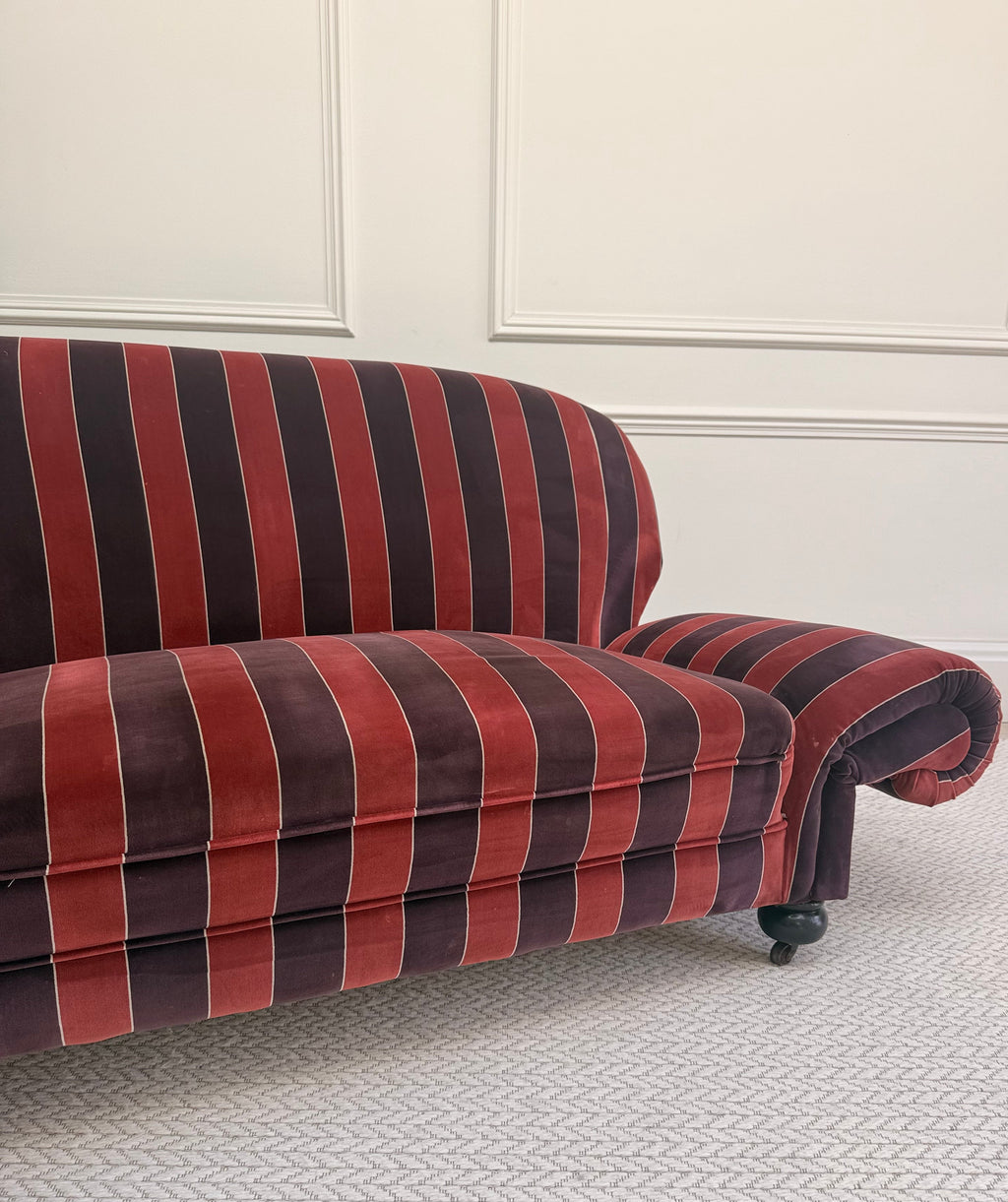 A close-up of a striped velvet loveseat sofa in red and dark brown with thin cream vertical stripes, set against a white paneled wall and textured light gray carpet.